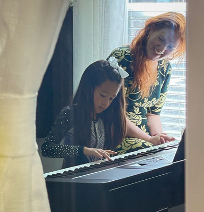 female piano teacher sits at piano with young student, smiling