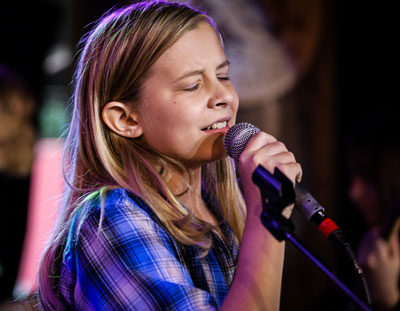 young boy with long blonde hair sings expressively into a microphone on stage