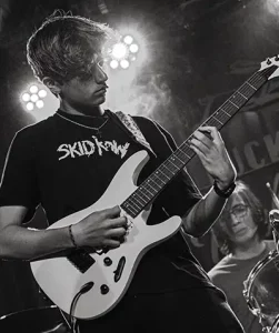 Tall teen boy plays rock guitar with drummer behind him on stage