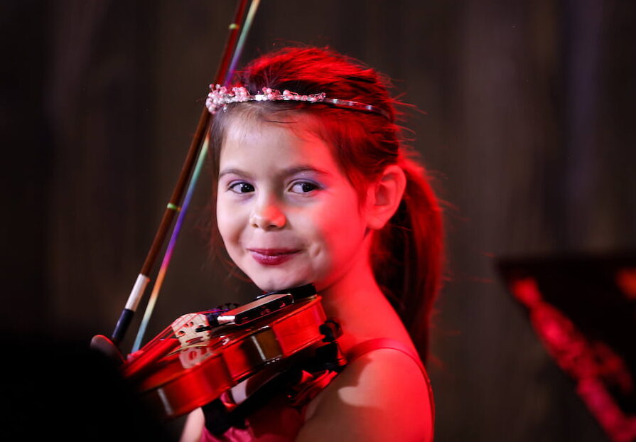 Young girl smiles at camera while playing a tiny violin in a recital